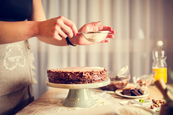 House wife wearing apron making finishing touches on birthday dessert chocolate cake.Woman making homemade cake with easy recipe,sprinkling powdered sugar on top Стоковое Фото